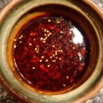 Close-up of a rich, dark red dumpling sauce with visible chili flakes and sesame seeds in a textured bowl.