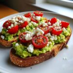 Two slices of avocado toast with feta cheese and halved cherry tomatoes, seasoned with pepper.