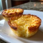 Two golden-brown baby lemon impossible pies on a white plate, one with a bite taken out.