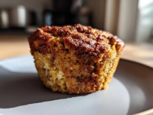 Close-up of a golden-brown banana protein muffin on a gray plate, bathed in sunlight.
