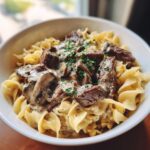 A close-up of a bowl of creamy Beef Stroganoff served over wide egg noodles, garnished with fresh parsley.