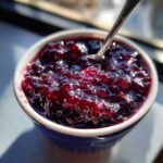 Close-up of glistening homemade blueberry jam in a blue and white bowl, with a spoon resting inside.