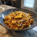 A close-up of a bowl filled with Chicken Pad Thai, featuring noodles, chicken, scrambled egg, bean sprouts, and crushed peanuts.