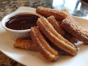 A plate of golden brown, sugar-cinnamon coated churros served with a small bowl of rich chocolate dipping sauce.