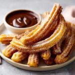 A plate of freshly fried churros coated in cinnamon sugar, served with a bowl of rich chocolate dipping sauce.