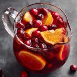 A clear glass pitcher filled with vibrant Cranberry Orange Punch, garnished with fresh cranberries and orange slices.