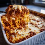 A serving spoon lifts a generous portion of creamy baked spaghetti casserole, showcasing melted cheese and rich meat sauce.
