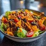 A close-up of a bowl of Dorito Taco Salad, featuring lettuce, seasoned ground beef, cherry tomatoes, black olives, and crushed Doritos, drizzled with dressing.