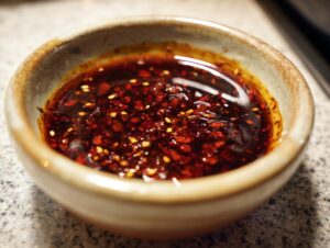 A close-up shot of a small bowl filled with a rich, dark red dumpling sauce, speckled with chili flakes and sesame seeds.