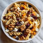 A close-up overhead view of a bowl of fall pasta salad, featuring fusilli pasta, butternut squash, cranberries, walnuts, and feta cheese.
