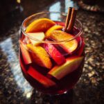 A glass of red fall sangria filled with slices of apples, oranges, and cinnamon sticks.