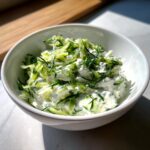 A close-up of creamy Tzatziki sauce in a white bowl, topped with shredded cucumber and fresh herbs.