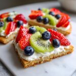 Close-up of delicious fruit pizza sugar cookie slices topped with cream cheese frosting and fresh strawberries, kiwi, and blueberries.