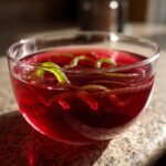 A clear glass bowl filled with vibrant red Halloween poison apple punch, garnished with green gummy worms.