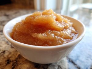 A close-up of a white bowl filled with warm, chunky homemade applesauce, showing its texture and cinnamon flecks.