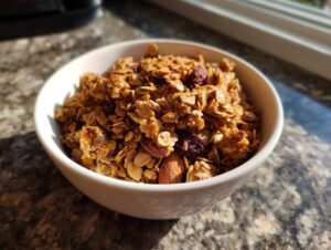 A white bowl filled with golden brown homemade granola, featuring oats, dried cranberries, and almonds.