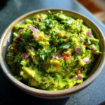 A close-up of a bowl of vibrant green Homemade Guacamole, topped with chopped red onion and fresh cilantro.