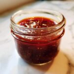 A close-up of a small glass jar filled with glistening, rich honey BBQ sauce.