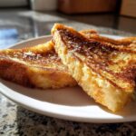 Close-up of golden brown protein French toast slices dusted with powdered sugar on a white plate.