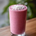 A tall glass filled with a vibrant pink smoothie, topped with shredded coconut. This is the Spooky Cinnamon Rolls Smoothie.