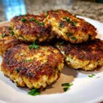 A close-up of a pile of golden-brown, freshly cooked tuna patties, garnished with chopped parsley.