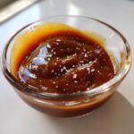 A close-up of thick, glossy salted caramel sauce topped with visible flakes of sea salt in a small glass bowl.