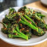 A close-up of bright green roasted broccoli topped with crispy, browned seasonings served on a white plate.