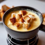 Close-up of creamy cheese fondue in a dark pot topped with golden toasted bread cubes.