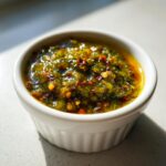 Close-up of chunky, vibrant green tomato salsa topped with red pepper flakes in a small white bowl.