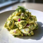 A mound of creamy avocado egg salad mixed with red onion and topped with fresh dill on a white plate.