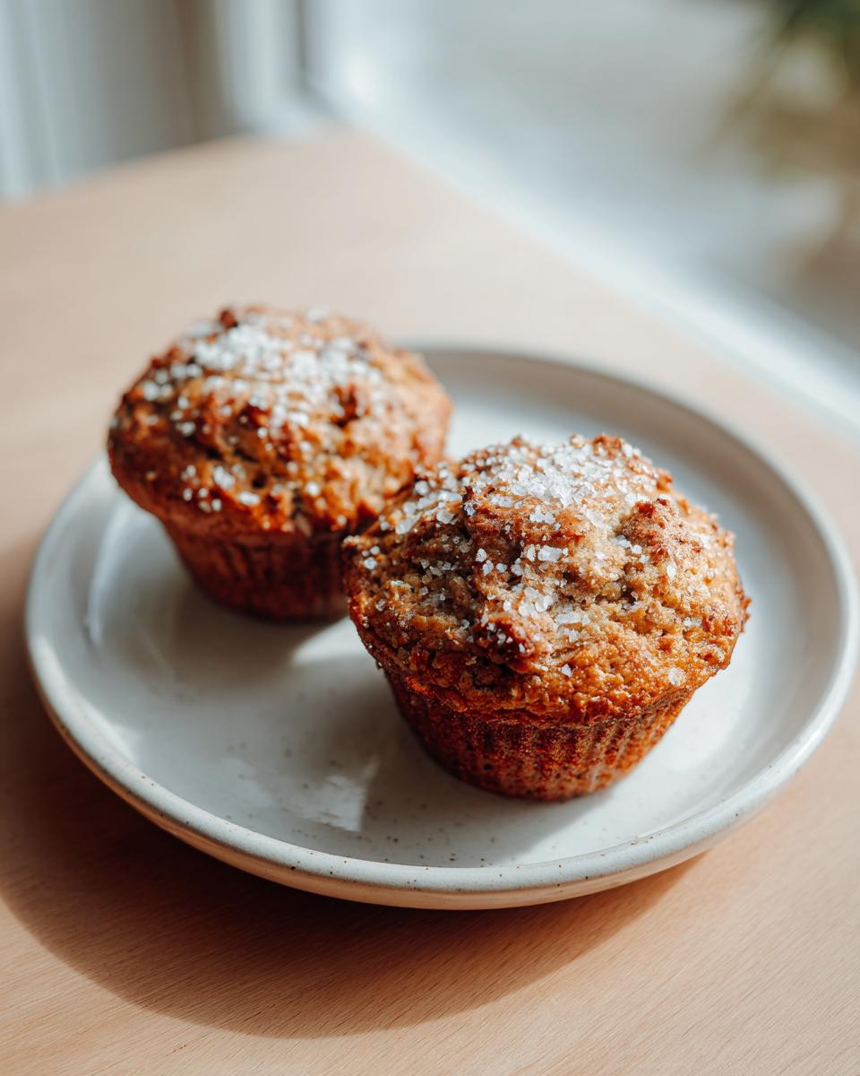 Two freshly baked banana oatmeal muffins topped with coarse sugar crystals, resting on a speckled white plate.