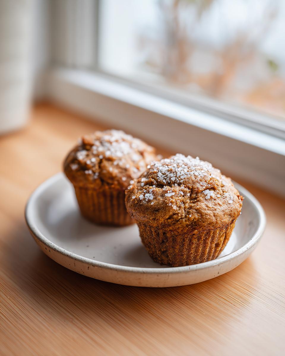 Two freshly baked banana oatmeal muffins topped with coarse sugar crystals, resting on a small white plate.