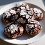 A pile of rich, dark chocolate crinkle cookies heavily dusted with white powdered sugar on a white plate.