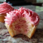 Close-up of a strawberry cupcake with a bite taken out, showing pink strawberry cake and bright pink frosting topped with freeze-dried strawberry pieces.