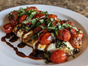 Plated grilled caprese chicken topped with melted mozzarella, roasted cherry tomatoes, fresh basil, and balsamic glaze.