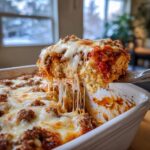 A serving spoon lifts a cheesy slice from a baked pizza casserole, showing long mozzarella cheese pulls.