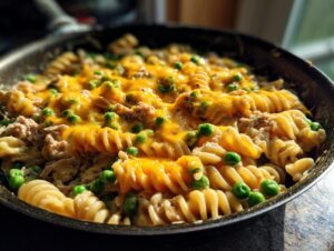 Close-up of creamy tuna pasta made with rotini, green peas, and melted cheddar cheese in a dark skillet.