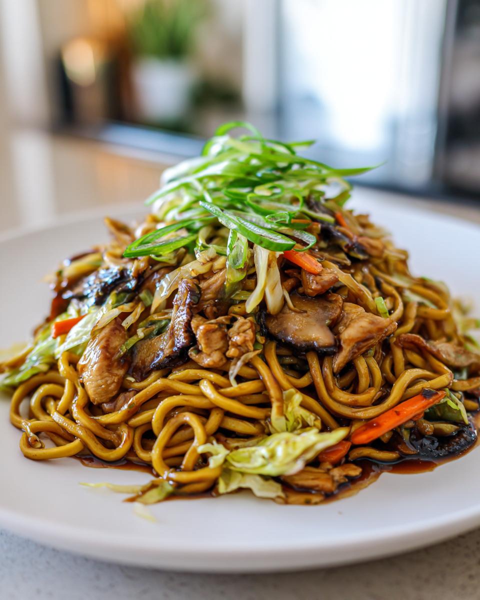 A close-up of a generous serving of chicken chow mein recipe noodles, mixed with vegetables and topped with fresh scallions.