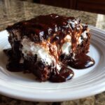 A rich slice of chocolate poke cake topped with white filling and dripping with glossy chocolate fudge sauce.