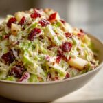 A close-up shot of creamy apple cranberry coleslaw featuring shredded cabbage, chunks of apple, and dried cranberries.