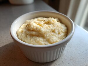 A close-up of creamy, yellow cheese grits served in a small white fluted ramekin, lightly seasoned.