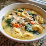A close-up of a white bowl filled with creamy chicken tortellini soup, featuring tortellini, shredded chicken, spinach, and carrots.