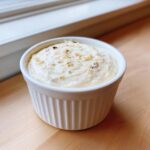 A small white ramekin filled with thick, creamy garlic sauce, topped with minced herbs or spices.