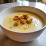 A close-up of a bowl of creamy, light-colored garlic soup topped with golden croutons and olive oil.