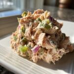 A mound of creamy salmon salad mixed with chopped green celery and purple red onion served on a white plate.