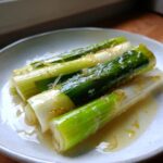 Close-up of three tender braised leeks, glistening with a light sauce, served on a simple white dish.