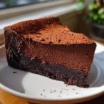 A rich, two-layered slice of flourless chocolate torte dusted generously with cocoa powder, sitting on a white plate.