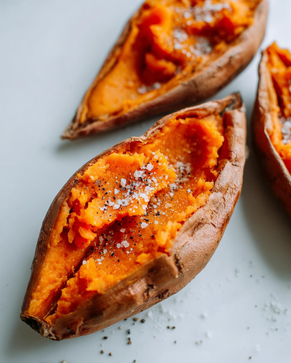 Close-up of fluffy baked sweet potatoes split open and seasoned with coarse salt and black pepper.