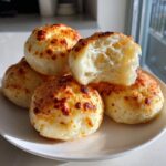 A stack of golden-brown brazilian cheese bread on a white plate, one piece broken open showing a chewy, airy interior.