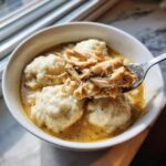 A spoonful of shredded chicken lifted from a bowl of fluffy chicken and dumplings in a savory, creamy broth.